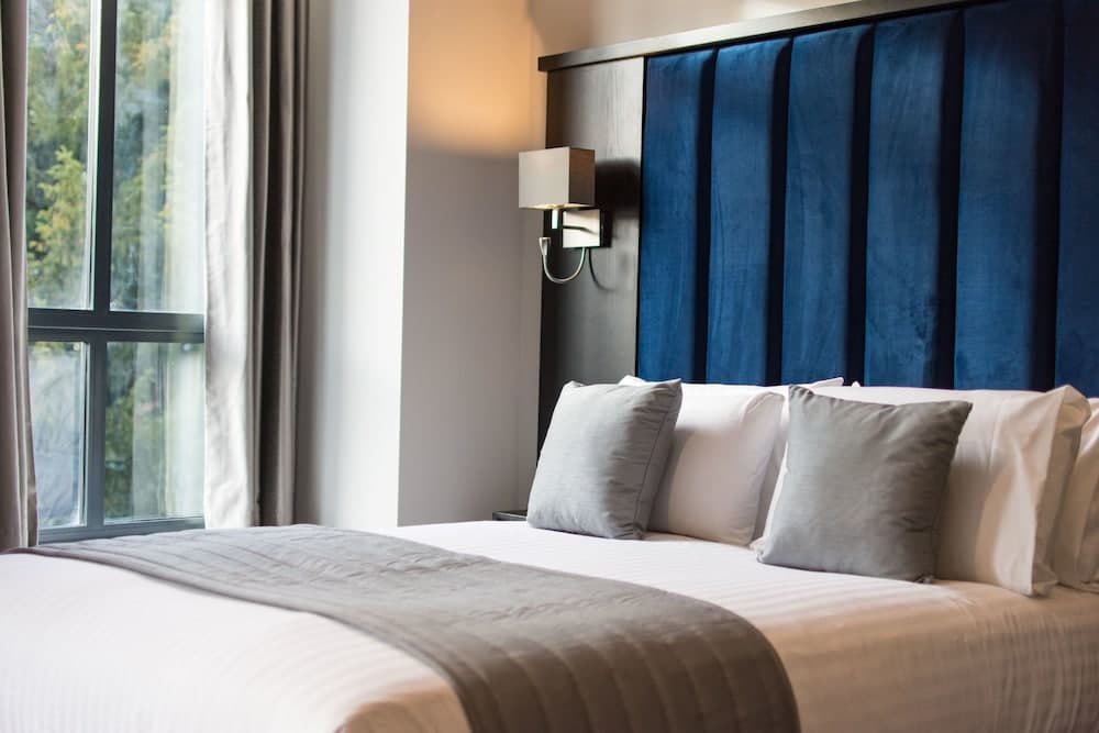 A cozy hotel room features a neatly made bed with a plush blue headboard, soft gray pillows, and a large window offering a serene view of greenery, ideal for travelers seeking relaxation and comfort.