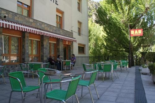 A serene outdoor café scene features green chairs and tables under striped awnings beside a stone building, set against lush greenery, ideal for travelers seeking a relaxing spot to unwind.
