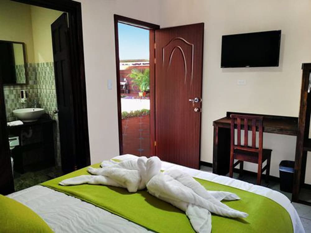 A cozy hotel room features a neatly made bed adorned with towel art, a desk with a chair, a mounted TV, and an open door leading to a sunny outdoor view, ideal for travelers seeking comfort and relaxation.
