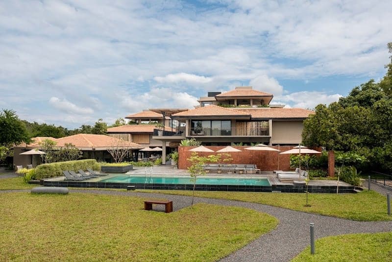 A serene tropical resort features a lush green lawn, a sparkling pool, and modern villas under a partly cloudy sky, ideal for luxury beachside relaxation in Goa, India.
