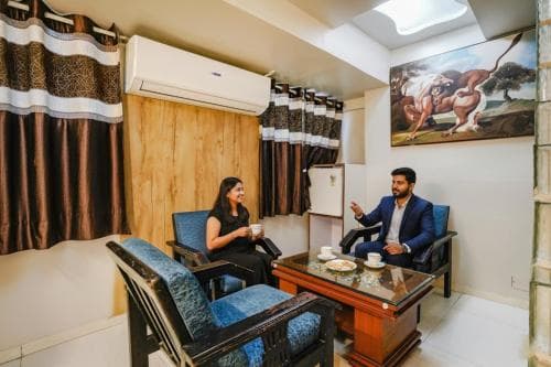 A cozy indoor lounge features two individuals enjoying tea amidst wooden furnishings, patterned curtains, and an artistic wall painting, ideal for travelers seeking a relaxed meeting or rest spot.