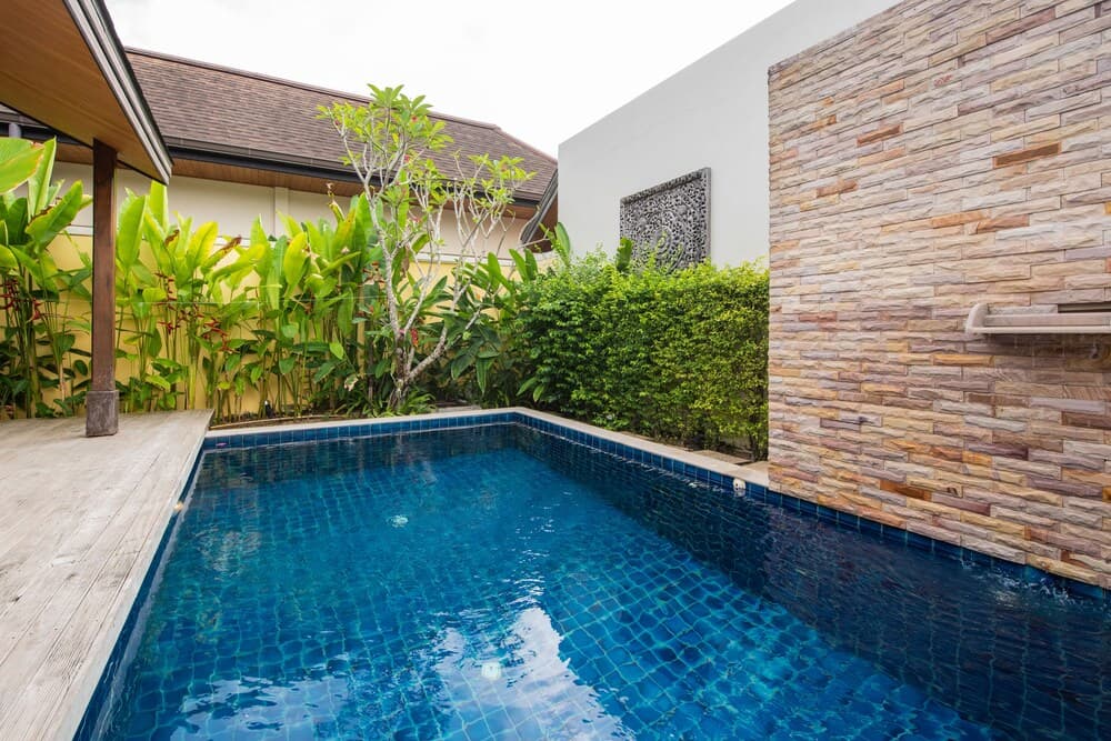 A serene tropical villa courtyard features a crystal-clear pool surrounded by lush greenery, a wooden deck, and a textured stone wall, ideal for a relaxing vacation retreat.