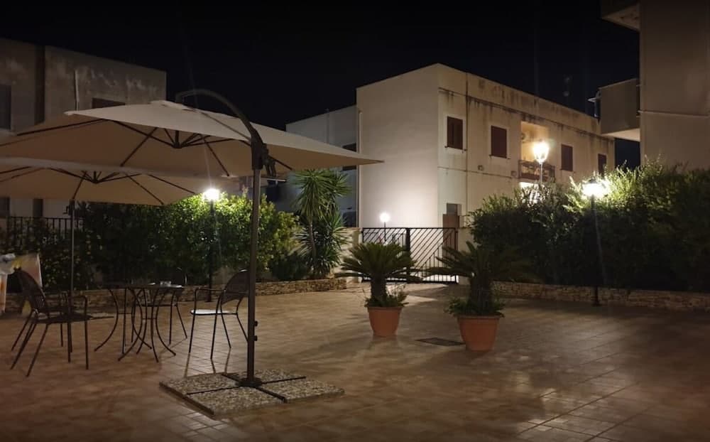 A serene nighttime courtyard illuminated by soft lighting features potted plants, outdoor seating under umbrellas, and a tranquil ambiance ideal for a relaxing evening retreat.