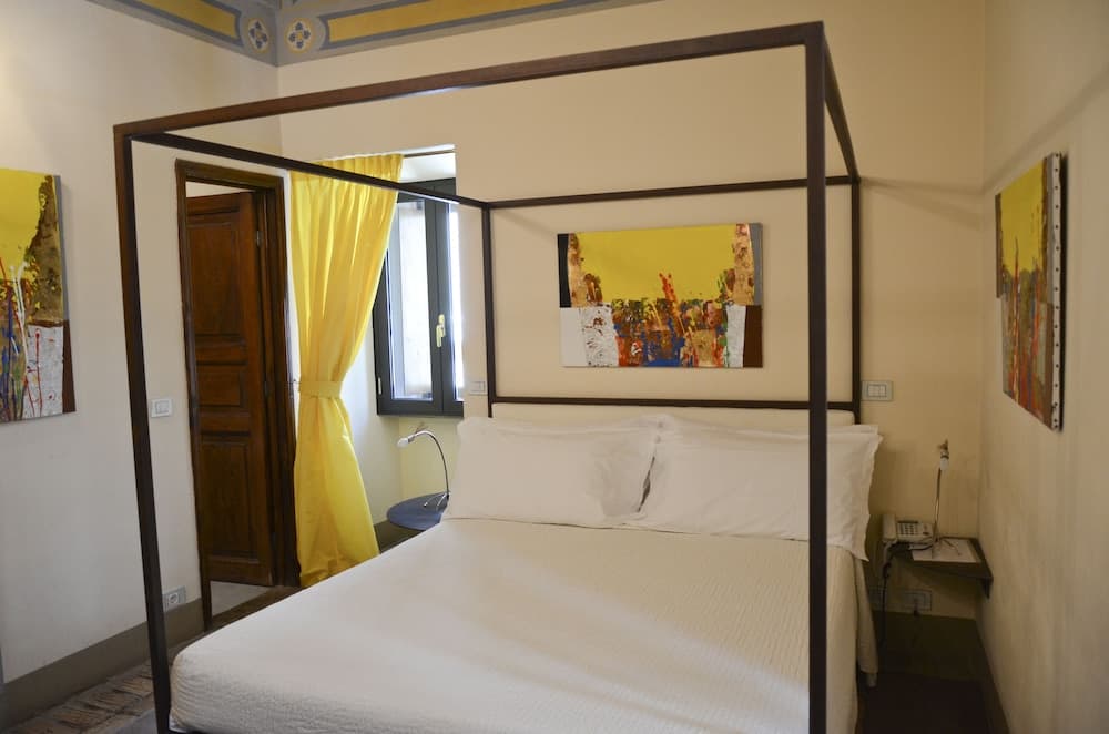A cozy hotel room features a four-poster bed adorned with white linens, vibrant yellow curtains, and abstract artwork, creating a warm and inviting atmosphere ideal for travelers seeking comfort and charm.
