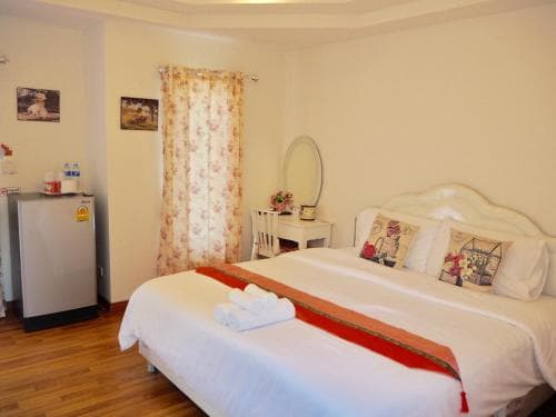 A cozy hotel room features a neatly made bed with red accents, floral curtains, a small fridge, and a mirror on a wooden desk, ideal for travelers seeking comfort in a budget-friendly accommodation.