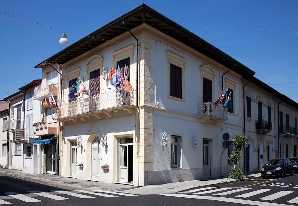 A charming European hotel with balconies adorned by colorful flags stands invitingly on a sunny street corner, perfect for travelers seeking a quaint and cozy accommodation experience.