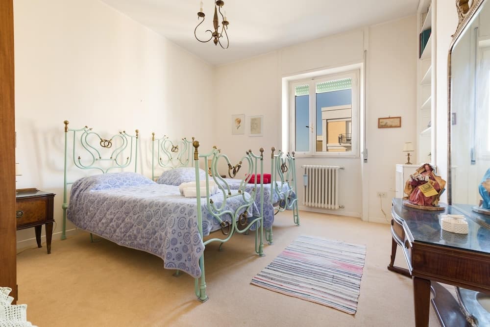 A cozy, vintage-style bedroom with twin beds framed in ornate green metal, soft carpeting, and a striped rug, exudes warmth and charm, ideal for travelers seeking a quaint and comfortable stay.