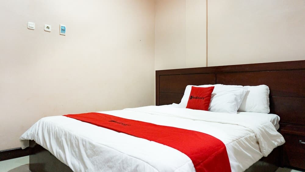 A cozy hotel room features a neatly made bed with white linens and a red accent pillow, set against neutral walls, ideal for travelers seeking comfort and simplicity.
