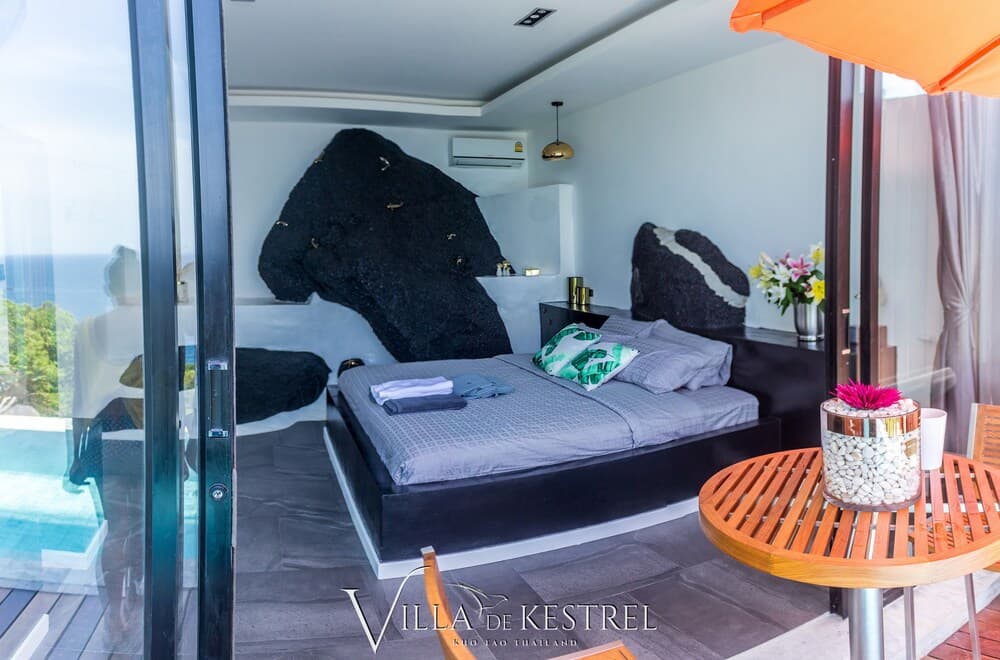 A serene bedroom with a modern design featuring large rock headboards overlooks the ocean through glass doors, ideal for travelers seeking luxury and tranquility in a tropical villa setting.