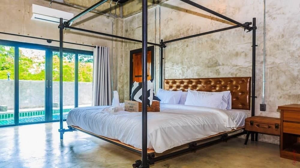A cozy industrial-style bedroom with exposed concrete walls, a metal canopy bed, and a view of lush greenery through large glass doors, ideal for travelers seeking a unique and serene accommodation experience.