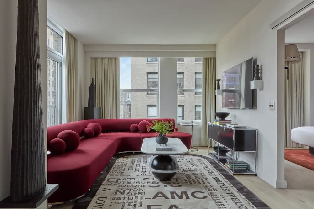 A modern urban apartment living room features a vibrant red sectional sofa, a stylish rug with text patterns, and large windows offering a cityscape view, ideal for travelers seeking a cozy yet sophisticated stay in a bustling metropolis.