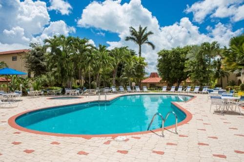 A serene tropical resort pool area surrounded by lush palm trees under a bright blue sky, featuring lounge chairs and tables, ideal for vacation relaxation.