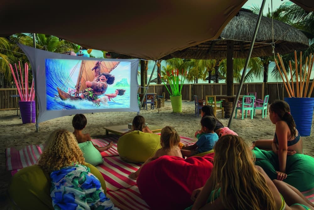 A lively beachside outdoor cinema under a canopy features families enjoying an animated movie on a large screen, surrounded by colorful bean bags, vibrant planters, and tropical palm trees, perfect for a family-friendly vacation activity.