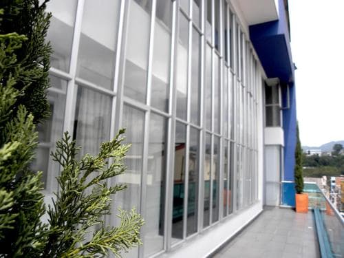 A serene balcony scene features a modern glass-paneled building with vibrant blue accents, lush greenery in the foreground, and distant hills under a clear sky, ideal for travelers seeking a tranquil urban retreat.