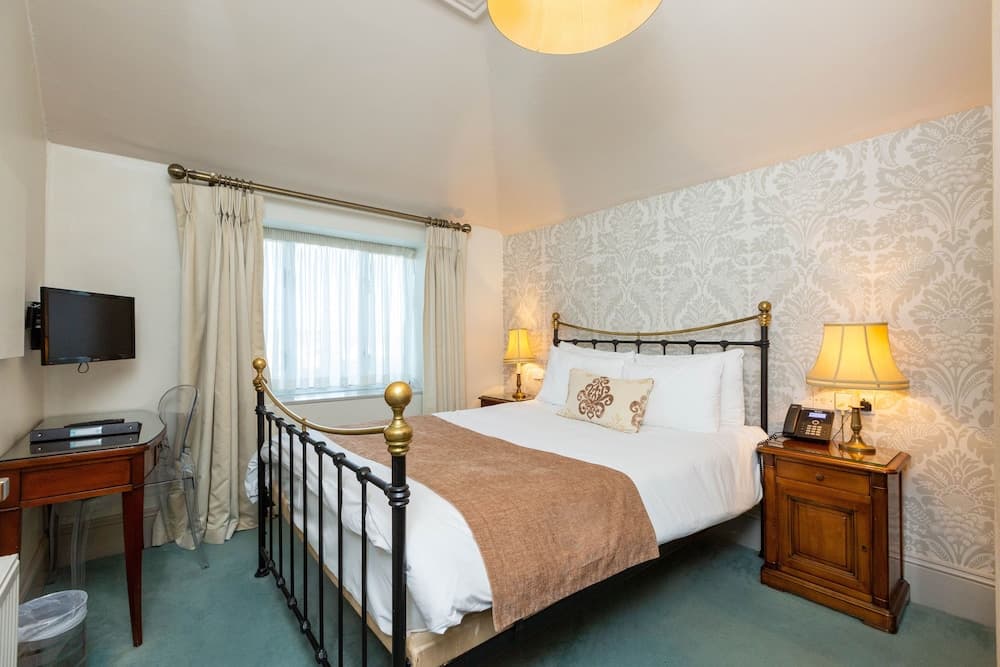 A cozy hotel room features a neatly made bed with a brass frame, patterned wallpaper, warm lighting from bedside lamps, and a desk with a TV, ideal for travelers seeking comfort and relaxation.