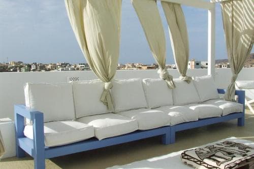 A serene rooftop lounge features a cozy blue sofa with white cushions under flowing beige curtains, overlooking a coastal town, ideal for travelers seeking relaxation and scenic views.