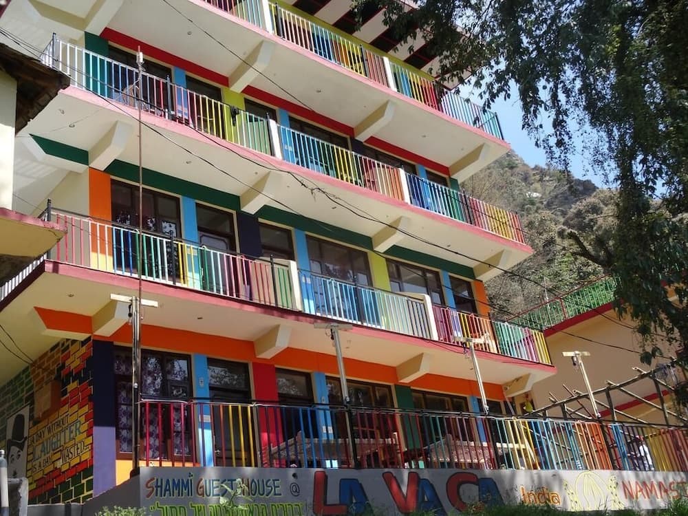A vibrant, colorful guesthouse with rainbow railings stands amidst lush greenery in a serene mountainous area, ideal for travelers seeking a cheerful and relaxing retreat in India.