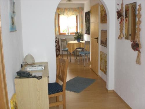 A cozy, warmly lit apartment hallway leads to a dining area with wooden furniture, floral decorations, and a bright window, ideal for travelers seeking a comfortable and homey stay.