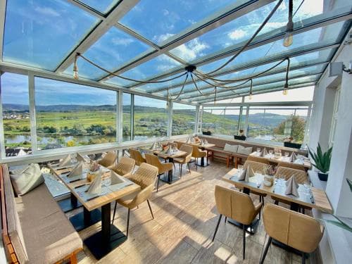 A bright, modern restaurant with a glass roof offers panoramic views of lush green landscapes and a river, creating an inviting atmosphere ideal for travelers seeking a relaxing dining experience with scenic beauty.