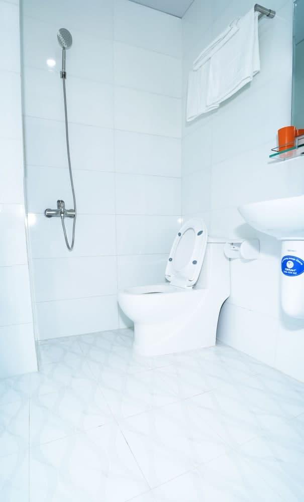 A clean, minimalist bathroom with white tiled walls and floors features a toilet, handheld shower, sink, and neatly hung towels, ideal for travelers seeking a simple yet functional accommodation space.