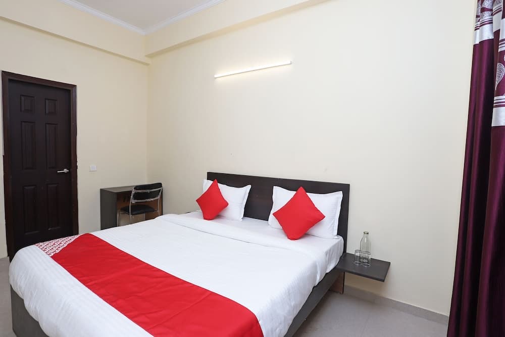 A cozy hotel room features a neatly made bed with red accents, a small desk by the door, and a minimalist design, ideal for travelers seeking comfort and simplicity.