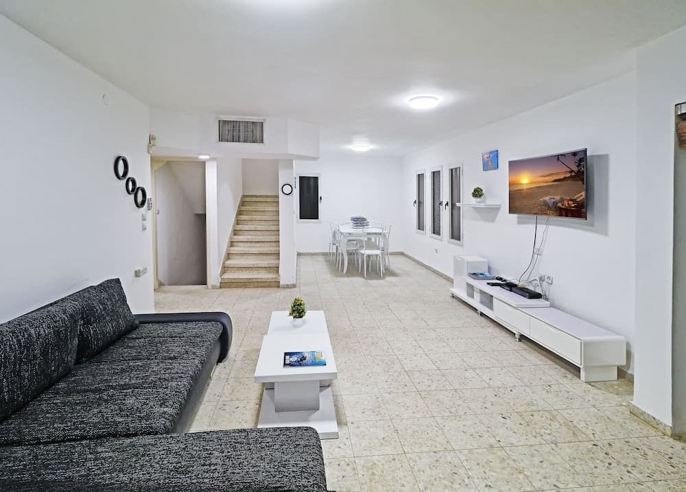 A bright, modern living room with white walls, tiled floors, a cozy sofa, dining area, staircase, and wall-mounted TV creates an inviting atmosphere ideal for vacation rentals or family stays.