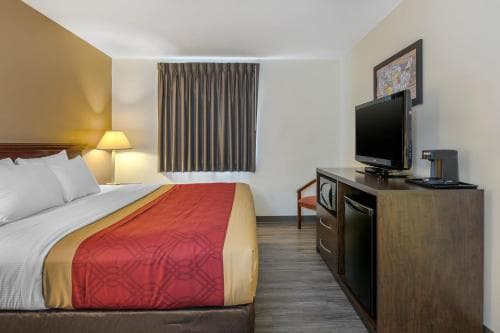 A cozy hotel room features a neatly made bed with red accents, a wooden dresser with a TV, and warm lighting, creating an inviting atmosphere ideal for travelers seeking comfort and relaxation.