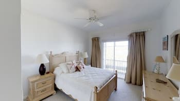 A serene bedroom with white walls, wooden furniture, and large windows draped in beige curtains offers a cozy retreat, ideal for travelers seeking relaxation and comfort.