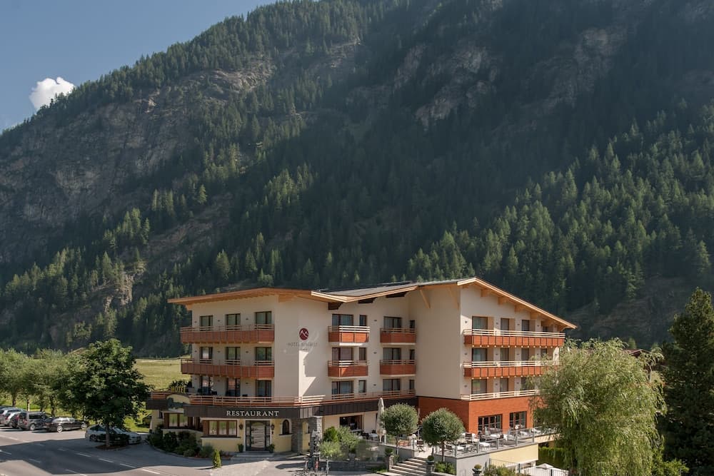 A serene mountain resort hotel with balconies and a restaurant sits nestled against lush green slopes, ideal for travelers seeking relaxation and scenic views in an alpine setting.