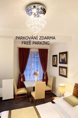 A cozy hotel room features a charming dining nook by the window, adorned with red curtains and framed artwork, illuminated by a sparkling chandelier, ideal for travelers seeking comfort with free parking amenities.