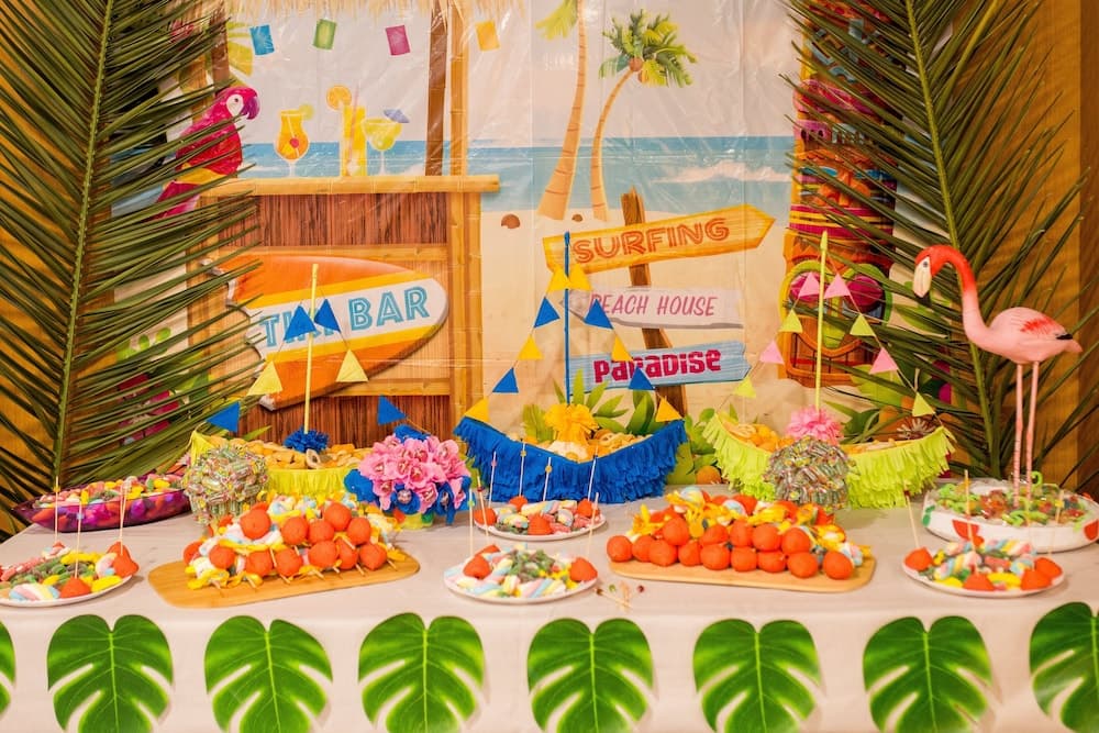 A vibrant tropical-themed party setup featuring colorful candy displays, palm leaves, flamingo decorations, and a "Surfing Paradise" backdrop creates a festive and inviting atmosphere perfect for a summer celebration or event planning inspiration.