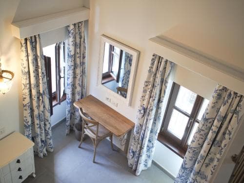 A cozy, well-lit hotel room features blue-flowered curtains framing wooden windows, a rustic wooden desk with a chair, and a mirror reflecting the serene ambiance, ideal for travelers seeking a charming and peaceful retreat.