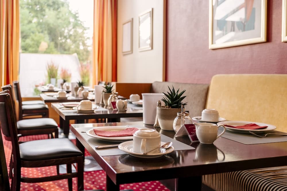 A cozy café with neatly set tables, vibrant orange curtains, and lush greenery outside offers a serene dining experience ideal for travelers seeking a relaxing meal spot.
