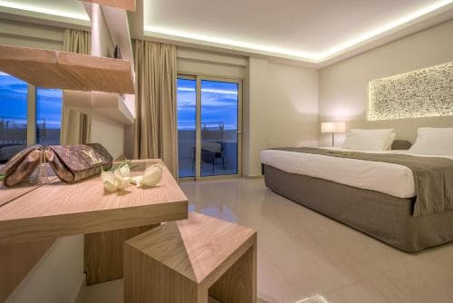 A serene hotel room with a modern design features a cozy bed, wooden furnishings, and a balcony overlooking the ocean at dusk, ideal for travelers seeking relaxation and comfort.
