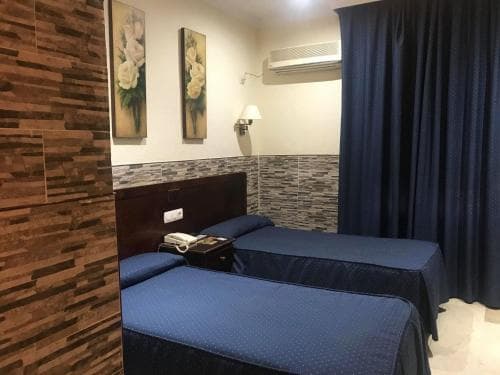 A cozy hotel room features two neatly made blue beds against a rustic wood-paneled wall, adorned with floral artwork, creating a welcoming atmosphere ideal for travelers seeking comfort and simplicity.