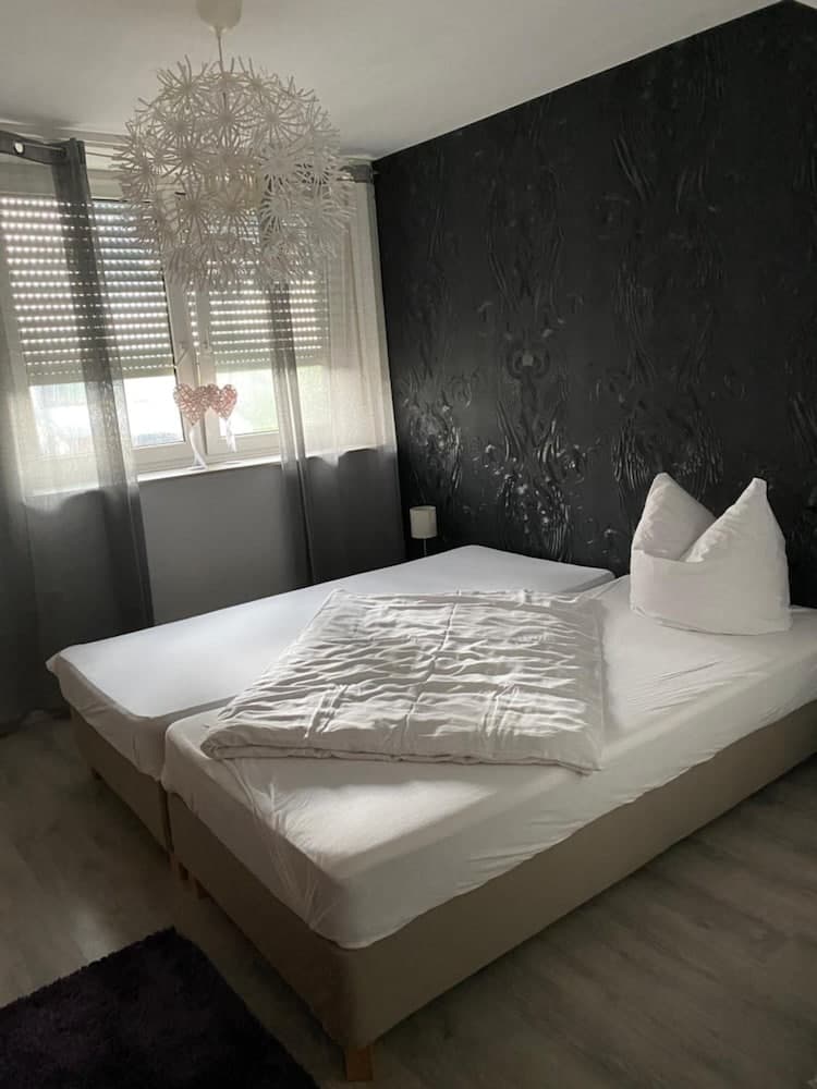 A cozy bedroom with a modern aesthetic features a large bed adorned in white linens, a textured black accent wall, and a unique dandelion-inspired chandelier, ideal for travelers seeking a stylish and serene accommodation.