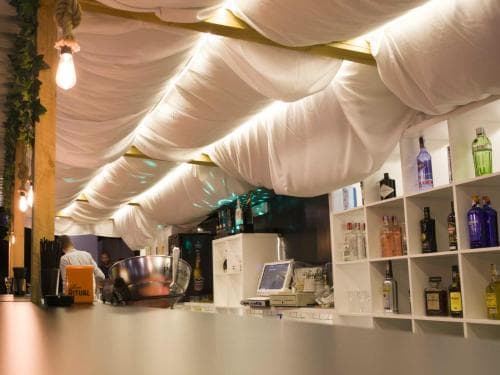 A cozy bar with draped white fabric ceilings, shelves lined with colorful bottles, and warm lighting creates an intimate atmosphere ideal for evening social gatherings.