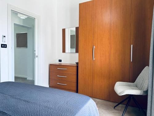 A cozy hotel room features a neatly made bed with blue bedding, wooden furniture including a wardrobe and dresser, a modern chair, and a mirror reflecting the serene ambiance, ideal for travelers seeking comfort and simplicity.