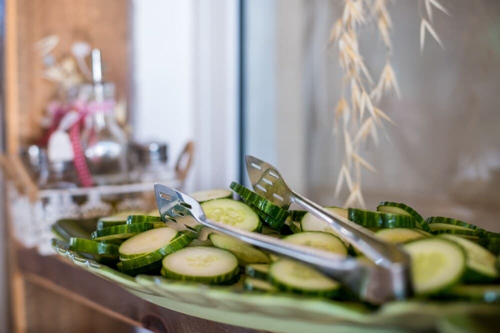 A cozy indoor setting features a plate of fresh cucumber slices with serving tongs, set against a rustic backdrop with decorative jars and dried wheat, ideal for a relaxing culinary retreat or home-cooked meal experience.
