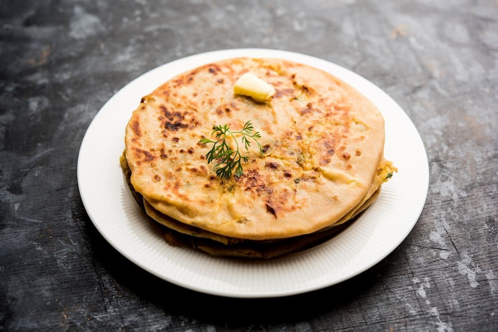 A stack of golden-brown parathas garnished with butter and herbs on a white plate against a rustic dark background, perfect for culinary enthusiasts exploring traditional Indian cuisine.