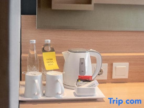 A cozy hotel room corner features a modern tea station with a white electric kettle, cups, and bottles adorned with an eco-friendly "Think Drink Save" note, ideal for travelers seeking convenience and sustainability during their stay.