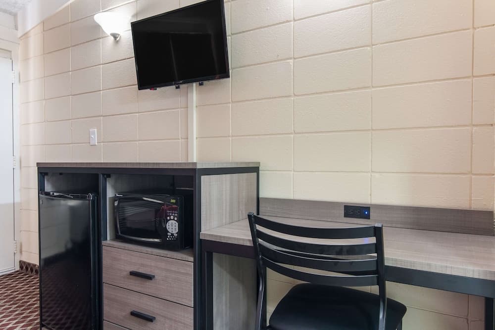 A cozy hotel room corner features a tiled wall backdrop with a mounted TV above a compact desk setup, including a microwave and mini-fridge, ideal for travelers seeking convenience and comfort.