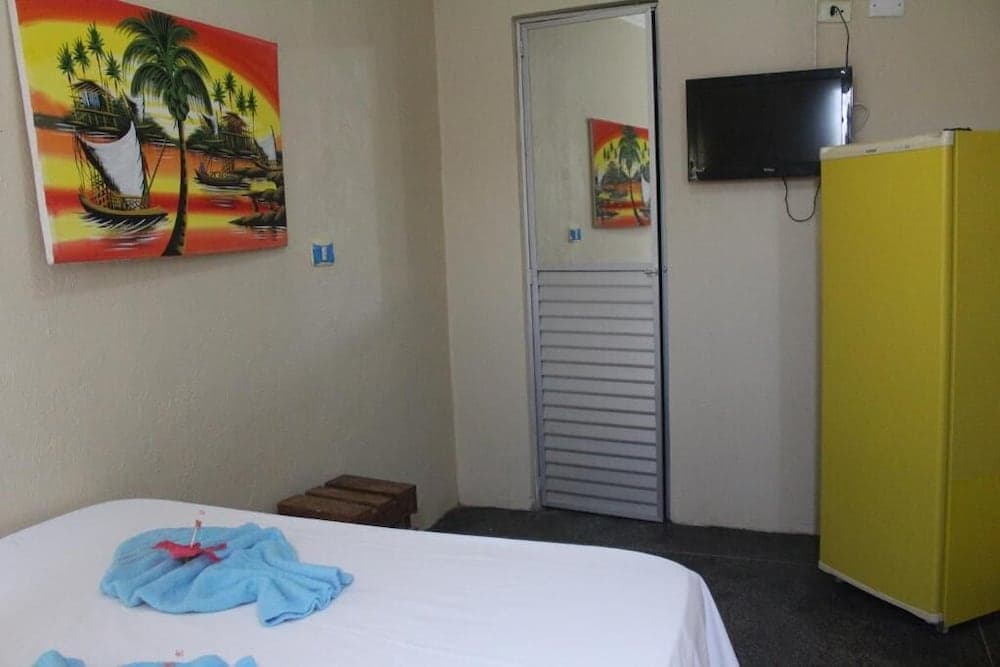 A cozy hotel room features a neatly made bed, vibrant tropical artwork, a yellow refrigerator, and a mounted TV, ideal for travelers seeking a comfortable and colorful retreat.