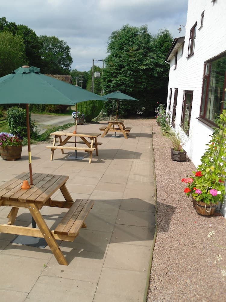 A cozy outdoor patio with wooden picnic tables under green umbrellas, surrounded by lush greenery and vibrant flower pots, offers a serene spot for travelers to relax and enjoy meals in a quaint countryside setting.