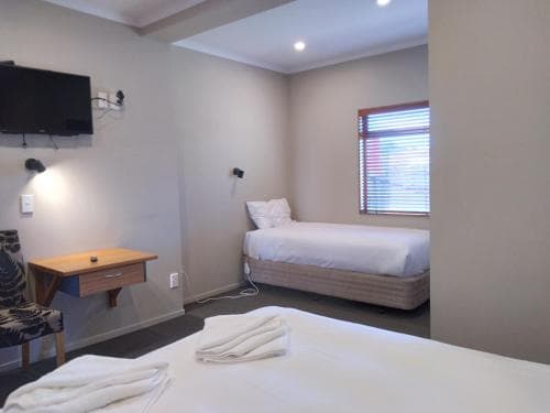 A cozy hotel room features a neatly made bed by a window with blinds, a mounted TV, a small desk, and soft lighting, ideal for travelers seeking comfort and simplicity.