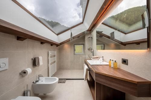 A serene attic bathroom with skylights framing mountain views offers a tranquil retreat, featuring modern fixtures, wooden accents, and ample natural light, ideal for luxury accommodation marketing.