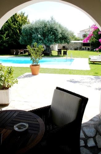 A serene outdoor patio features a cozy seating area under an archway, overlooking a tranquil pool surrounded by lush greenery and blooming flowers, ideal for a relaxing vacation retreat.