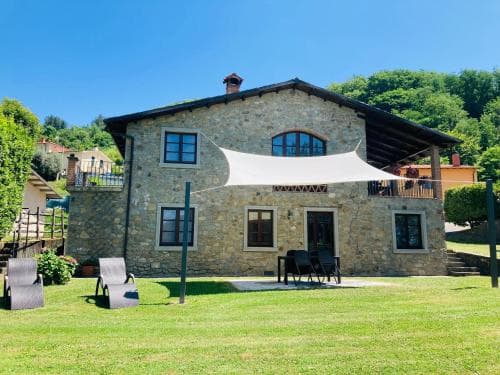 A serene stone villa with a lush green lawn, outdoor seating under a white canopy, and a picturesque hillside backdrop offers an idyllic retreat for travelers seeking relaxation in a countryside setting.