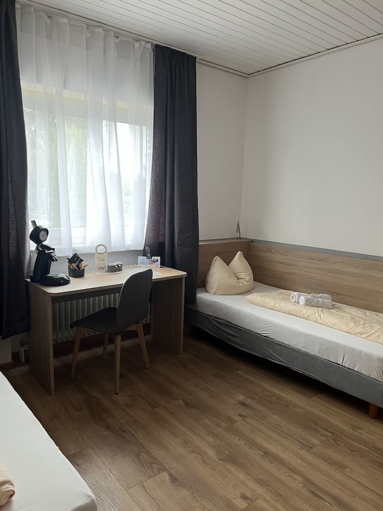 A cozy hotel room with wooden flooring, a neatly made bed adorned with pillows and towels, a desk featuring a coffee maker and chair by a window with sheer curtains, ideal for travelers seeking comfort and convenience.