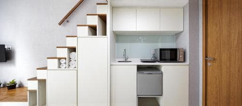 A cozy apartment kitchenette features sleek white cabinets, a compact dishwasher, and a staircase with built-in storage, creating a modern and functional space ideal for short-term rentals or city getaways.